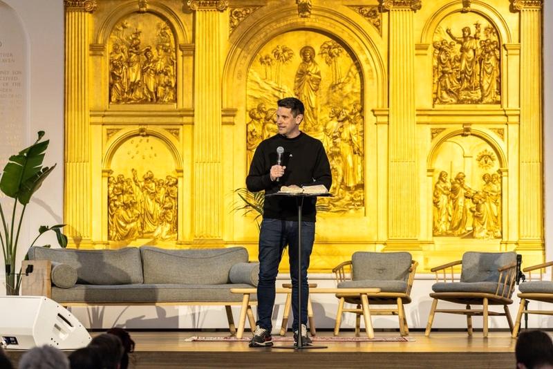 A speaker on stage in front of a gilded altarpiece, with sofas set out for a panel
